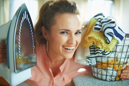 Portrait of smiling elegant 40 years old housewife in pink shirt and white pants with iron and...