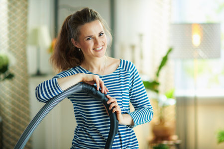 Portrait of happy young female in striped t-shirt and white pants with vacuum cleaner in the modern house in sunny day.の写真素材