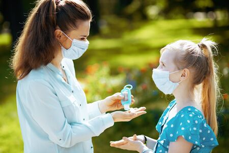 Life during coronavirus pandemic. stylish mother and daughter with medical mask disinfecting hands with sanitizer outdoors in the city park.の写真素材