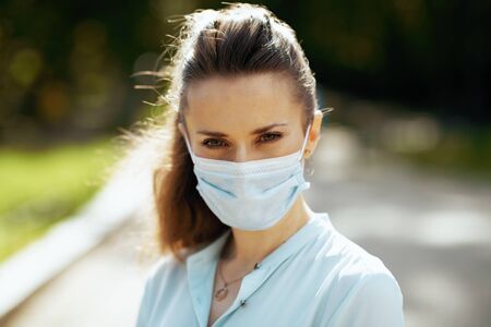Life during covid-19 pandemic. Portrait of young woman in blue blouse with medical mask outdoors in the city park.の写真素材