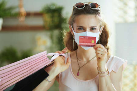Life during coronavirus pandemic. Portrait of modern woman shopper in pink blouse with medical mask and paper shopping bags talking on a smartphone.の写真素材