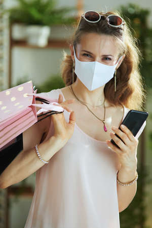 Life during coronavirus pandemic. Portrait of modern woman shopper in pink blouse with medical mask and paper shopping bags talking on a smartphone.の写真素材