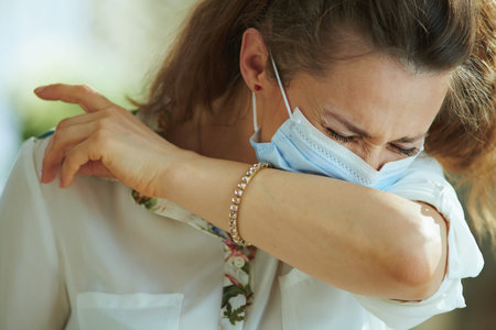 Life during covid-19 pandemic. trendy female in white blouse with medical mask sneezing into elbow.