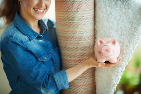 happy young housewife in jeans shirt and white pants with white carpet showing piggy bank in the modern living room in sunny day.の写真素材