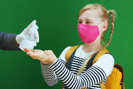 school girl with masks, yellow backpack and textbook disinfecting hands with sanitizer isolated on chalkboard green.の写真素材