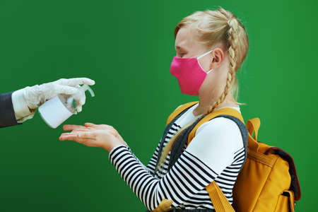 school girl with masks, yellow backpack and textbook disinfecting hands with sanitizer isolated on chalkboard green.の写真素材