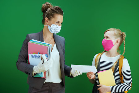 teacher woman and school girl with masks, yellow backpack and textbook isolated on chalkboard green.