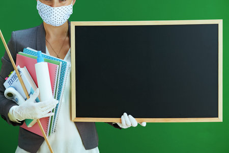 40 years old woman teacher in white blouse with medical mask, rubber gloves, digital thermometer, textbook and sanitizer isolated on chalkboard green background.の写真素材