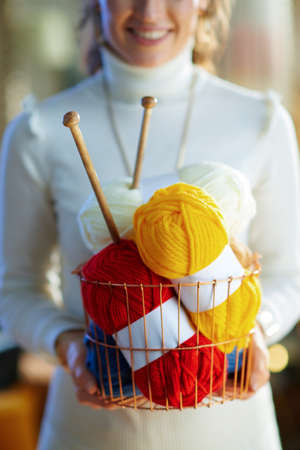 happy elegant middle age housewife in white sweater and skirt in the modern house in sunny winter day holding yellow and red knitting yarn hunks.の写真素材