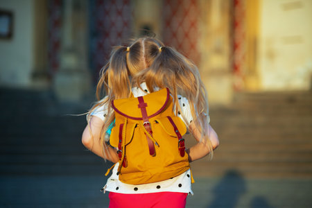 Life during covid-19 pandemic. Seen from behind girl in white polka dot blouse with yellow backpack going from school outside.の写真素材