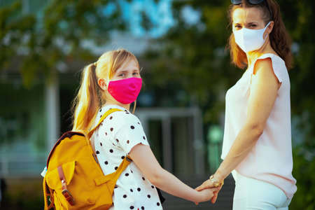 Life during coronavirus pandemic. young mother and daughter with masks and yellow backpack say goodbye before going to school outdoors.の写真素材