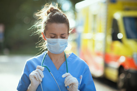 covid-19 pandemic. pensive modern medical doctor woman in scrubs with stethoscope and medical mask outside near ambulance.の写真素材