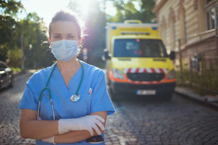 covid-19 pandemic. pensive modern medical doctor woman in scrubs with stethoscope and medical mask outside near ambulance.の写真素材