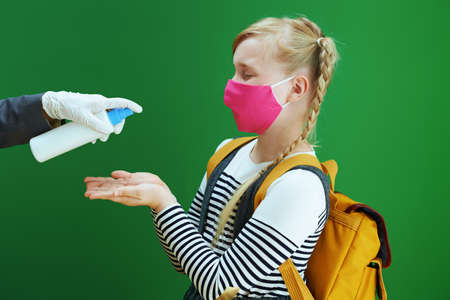 modern teacher woman and school girl with masks, yellow backpack and textbook measures temperature with digital thermometer isolated on chalkboard green.の写真素材