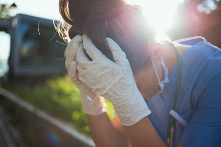 coronavirus pandemic. unhappy modern physician woman in scrubs with stethoscope and medical mask using a smartphone outdoors near clinic.の写真素材