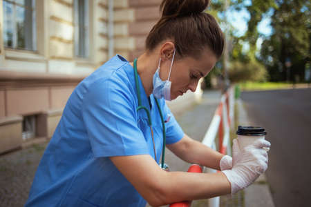 coronavirus pandemic. unhappy modern physician woman in scrubs with stethoscope and medical mask using a smartphone outdoors near clinic.の写真素材