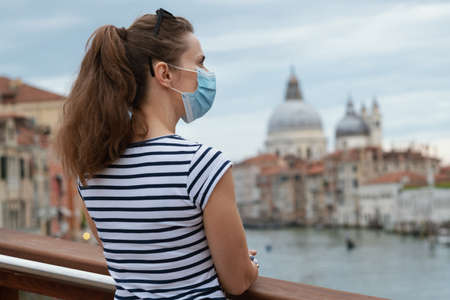 Travel during coronavirus pandemic. Seen from behind modern solo tourist woman with medical mask enjoying promenade on Accademia bridge in Venice, Italy.の写真素材