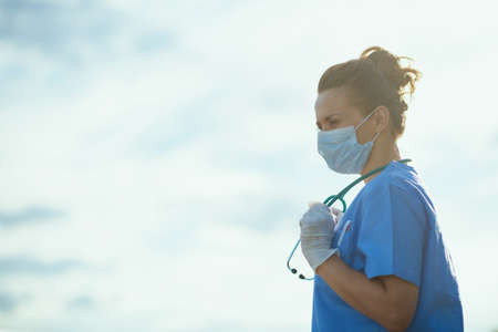 covid-19 pandemic. tired modern physician woman in scrubs with stethoscope, medical mask and rubber gloves outside near wall.の写真素材