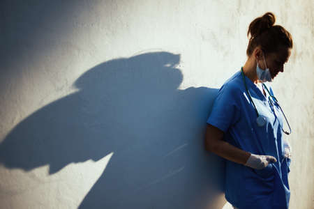 covid-19 pandemic. tired modern physician woman in scrubs with stethoscope, medical mask and rubber gloves outside near wall.の写真素材