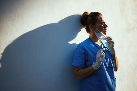 covid-19 pandemic. tired modern physician woman in scrubs with stethoscope, medical mask and rubber gloves outside near wall.の写真素材