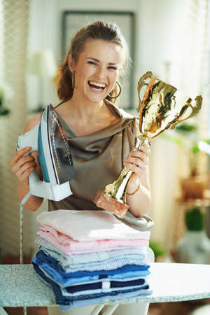 Closeup on happy woman in silk blouse and beige pants with steam iron, ironing board, pile of folded ironed clothes and gold goblet at home in sunny day.の写真素材
