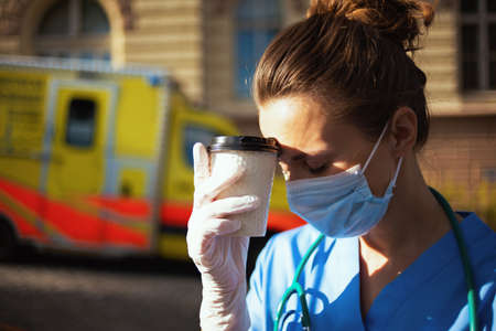covid-19 pandemic. tired modern paramedic woman in uniform with stethoscope, medical mask and cup of coffee outside near ambulance.の写真素材