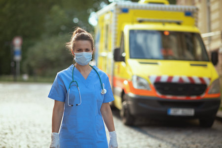 covid-19 pandemic. tired modern paramedic woman in uniform with stethoscope, medical mask and cup of coffee outside near ambulance.の写真素材