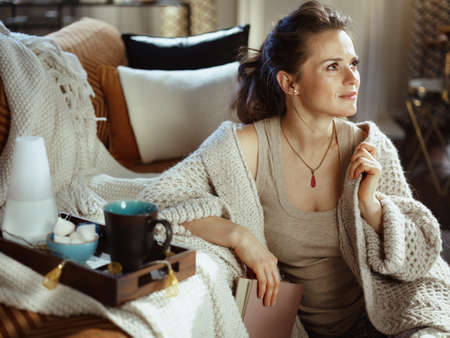 pensive young female in knitted cosy cardigan with book in the modern living room in sunny autumn day.の写真素材