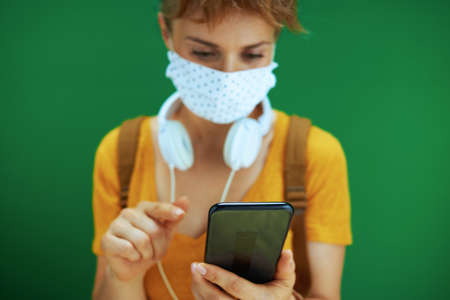 Life during covid-19 pandemic. Portrait of modern student woman in yellow shirt with textbooks, notebooks, medical mask, white headphones and backpack isolated on green.の写真素材
