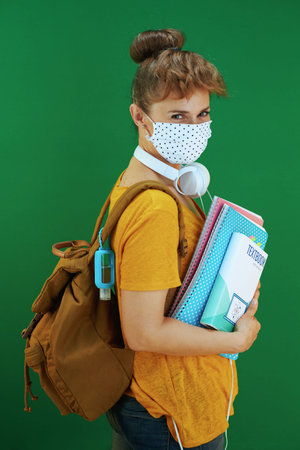 Life during covid-19 pandemic. Portrait of modern student woman in yellow shirt with textbooks, notebooks, medical mask, white headphones and backpack isolated on green.の写真素材