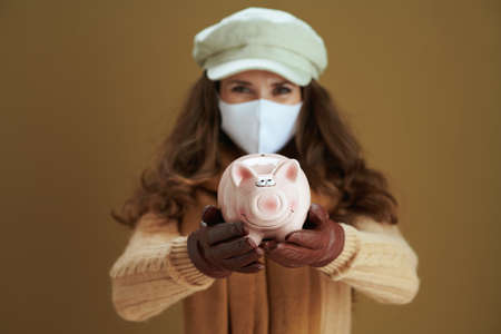 Life during coronavirus pandemic. cheerful elegant 40 years old housewife in scarf with white medical mask and yellow autumn maple leaves against beige background.の写真素材