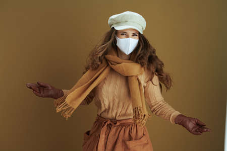 Life during coronavirus pandemic. cheerful elegant 40 years old housewife in scarf with white medical mask and yellow autumn maple leaves against beige background.の写真素材