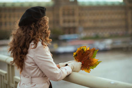 Hello november. happy modern middle aged woman in beige trench coat and black beret with shopping bags and autumn yellow leaves outdoors on the city street in autumn.の写真素材