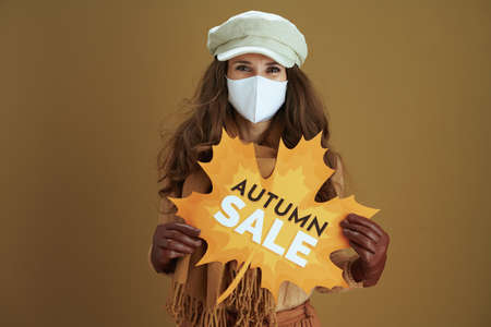 Life during coronavirus pandemic. smiling trendy 40 years old woman in sweater with white medical mask, yellow autumn maple leaves and paper shopping bag on brown background.の写真素材