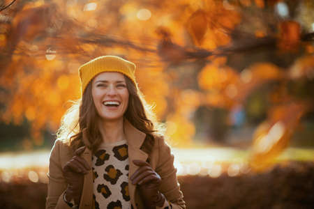 Hello autumn. smiling modern female in brown coat and yellow hat with autumn yellow leaves outdoors in the city park in autumn.の写真素材