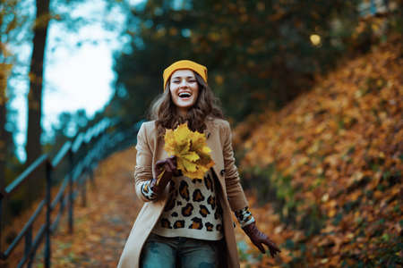 Hello autumn. smiling modern female in brown coat and yellow hat with autumn yellow leaves outdoors in the city park in autumn.の写真素材
