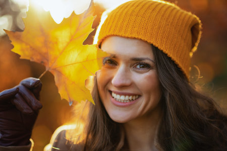 Hello autumn. smiling modern female in brown coat and yellow hat with autumn yellow leaves outdoors...