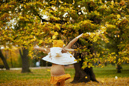 Seen from behind girl in brown sweater and orange hat rejoicing outdoors in the city park in autumn.の写真素材