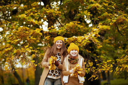 Hello autumn. Seen from behind modern mother and child in yellow hats with autumn yellow leaves enjoying promenade outside in the city park in autumn.の写真素材