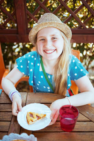 Portrait of smiling trendy girl in blue overall and hat with crostata sitting at the table having brunch in the terrace.の写真素材
