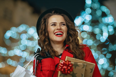 Winter Fun. smiling modern tourist woman in red coat and black beret with shopping bags, gifts and smartphone browsing online shop outside in the city street in the evening.の写真素材