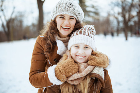 happy elegant mother and daughter in a knitted hats and sheepskin coats with mittens and medical mask in a knitted hat and sheepskin coat rejoicing outside in the city park in winter.の写真素材