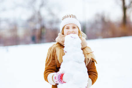 happy stylish girl in a knitted hat and sheepskin coat with mittens and snowman outdoors in the city park in winter.の写真素材