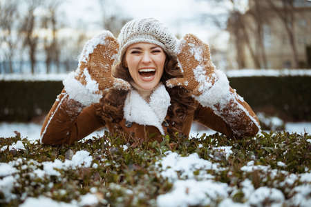 smiling stylish 40 years old woman with mittens in a knitted hat and sheepskin coat using smartphone app outside in the city park in winter.の写真素材
