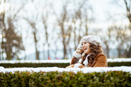 smiling stylish 40 years old woman with mittens in a knitted hat and sheepskin coat using smartphone app outside in the city park in winter.の写真素材