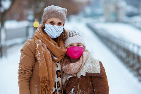 smiling elegant mother and child in a knitted hats and coats with medical mask walking outdoors in the city in winter.の写真素材