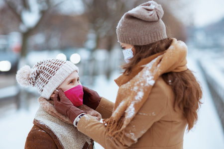 smiling elegant mother and child in a knitted hats and coats with medical mask walking outdoors in...