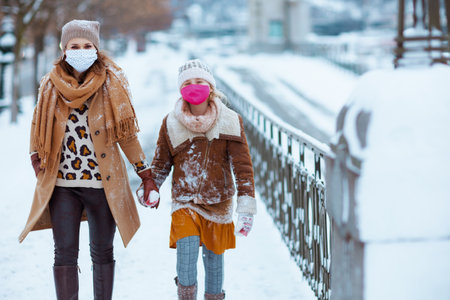 smiling elegant mother and child in a knitted hats and coats with medical mask walking outdoors in the city in winter.の写真素材