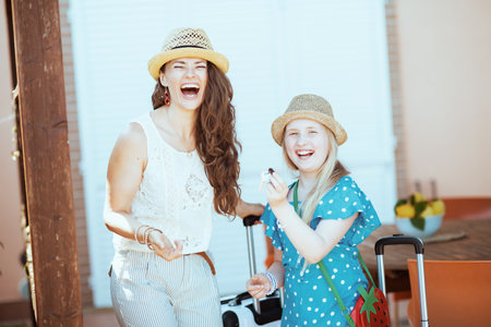 Seen from behind trendy mother and daughter tourists in hats with trolley bags in the terrace of guest house hotel.の写真素材