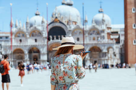 Seen from behind traveller woman in floral dress with hat speaking on a smartphone at San Marco square in Venice, Italy.の写真素材